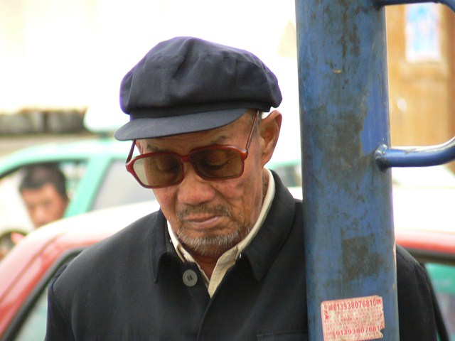 a man in a hat and sunglasses leans against a pole