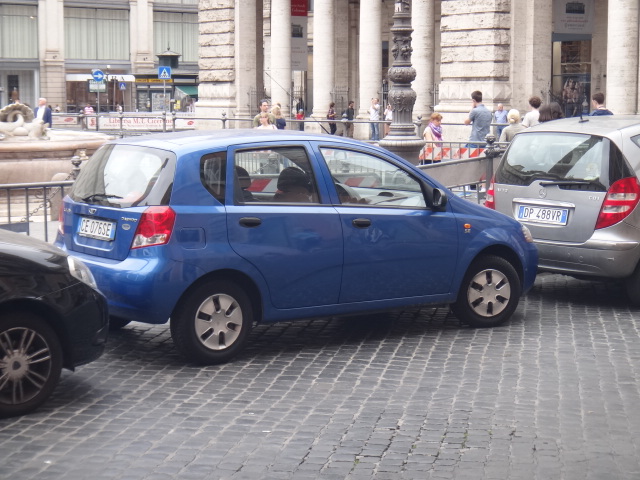a small blue car and two other cars in front of a very tall building