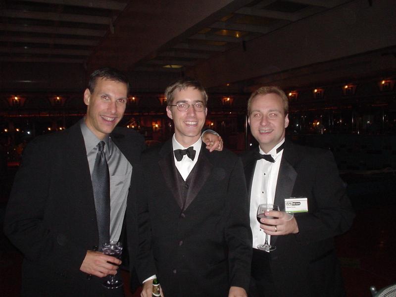 three men in tuxedos smile and stand smiling