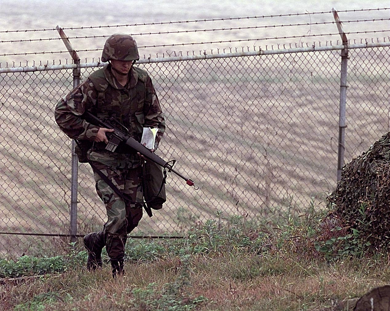 military men in camouflage, carrying guns and walking behind a barbed wire fence