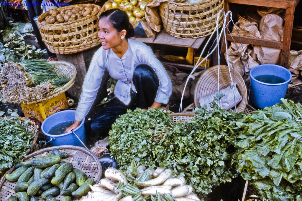 an asian woman crouches on baskets at a vegetable market
