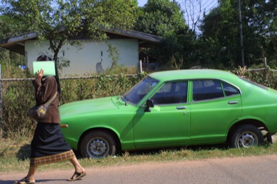 a lady walks past a green car with an old woman standing on it