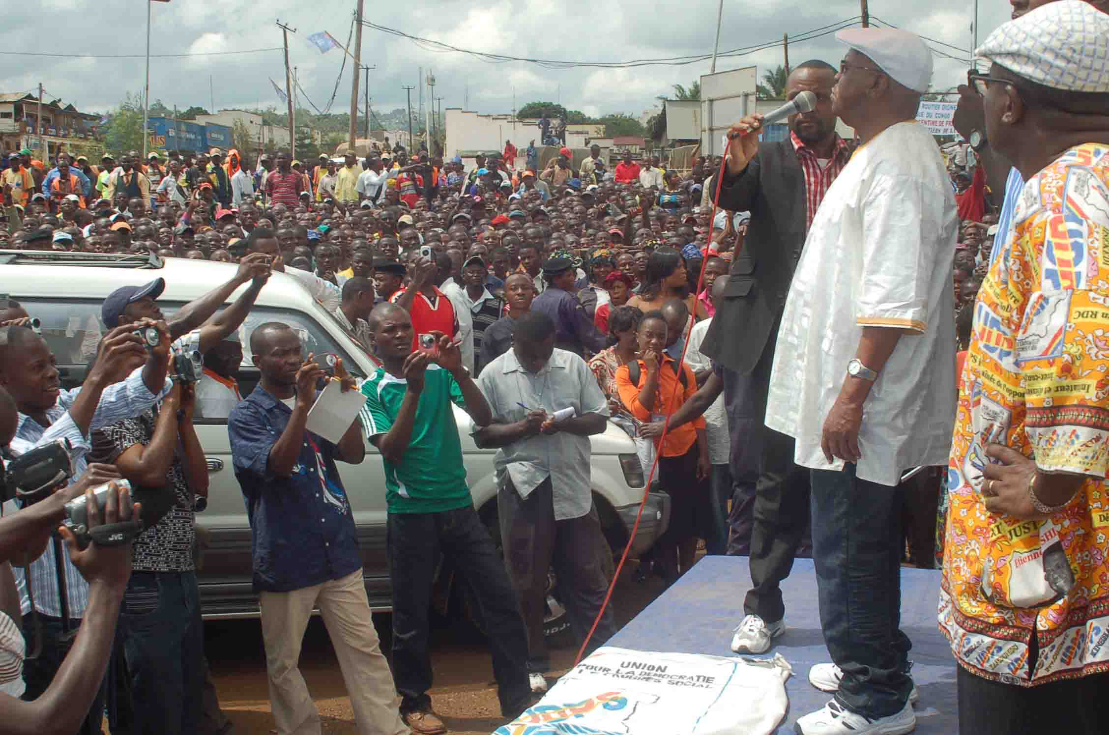 a man in white shirt standing next to car with crowd behind him