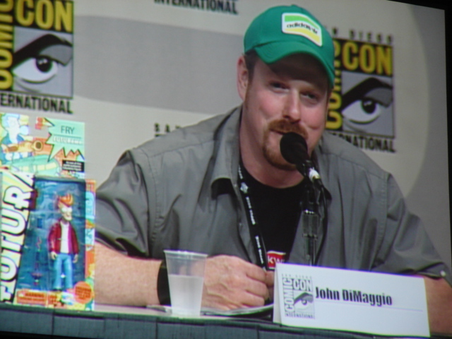 man with funny hat sitting at the microphone in front of comic convention booth