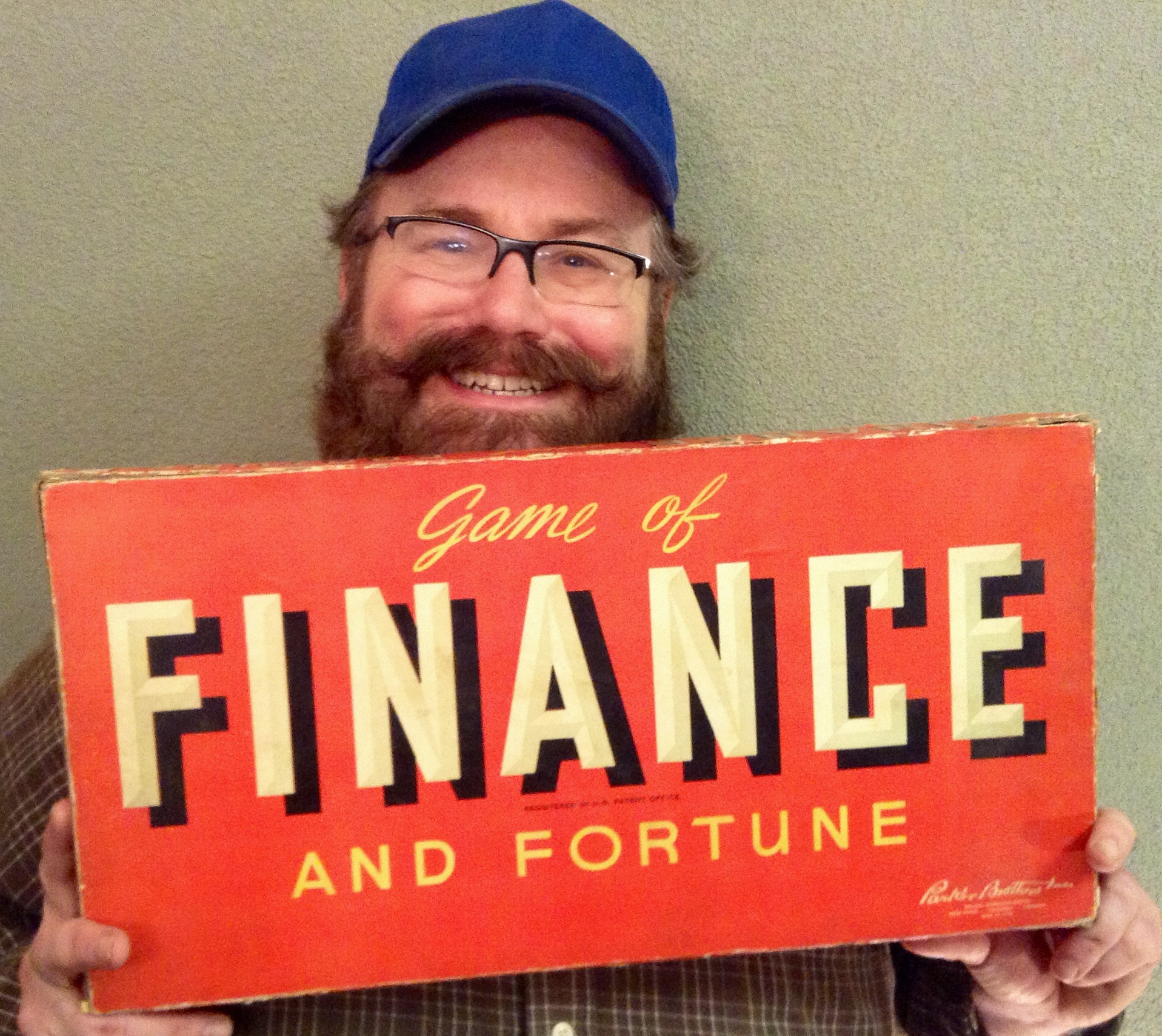 a bearded man smiles while holding a board game that has the words finance and fortune written on it