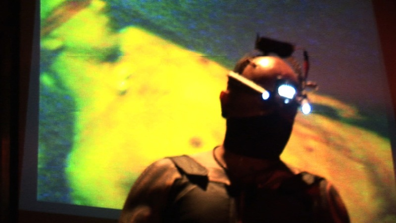 a man stands in front of a screen that is lit up with glowing eyes and a futuristic head