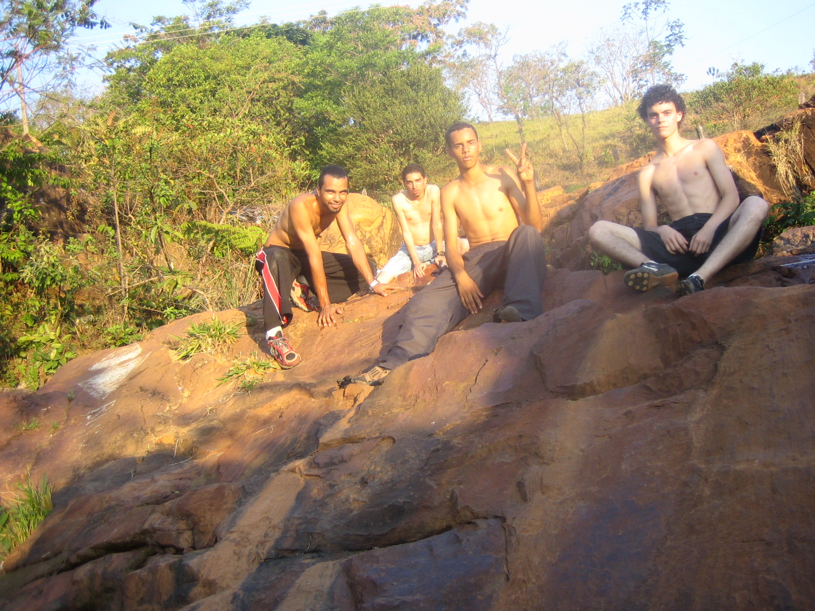five shirtless young men sit on large rocks as they watch someone climb a hill