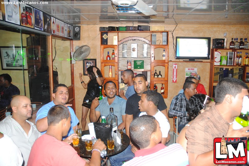 group of people drinking at a bar with the man standing up holding a beer bottle