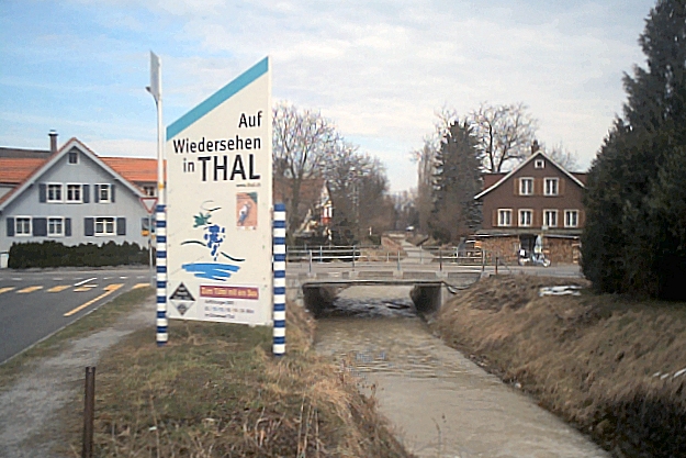 a welcome sign at a river side, with buildings in the background