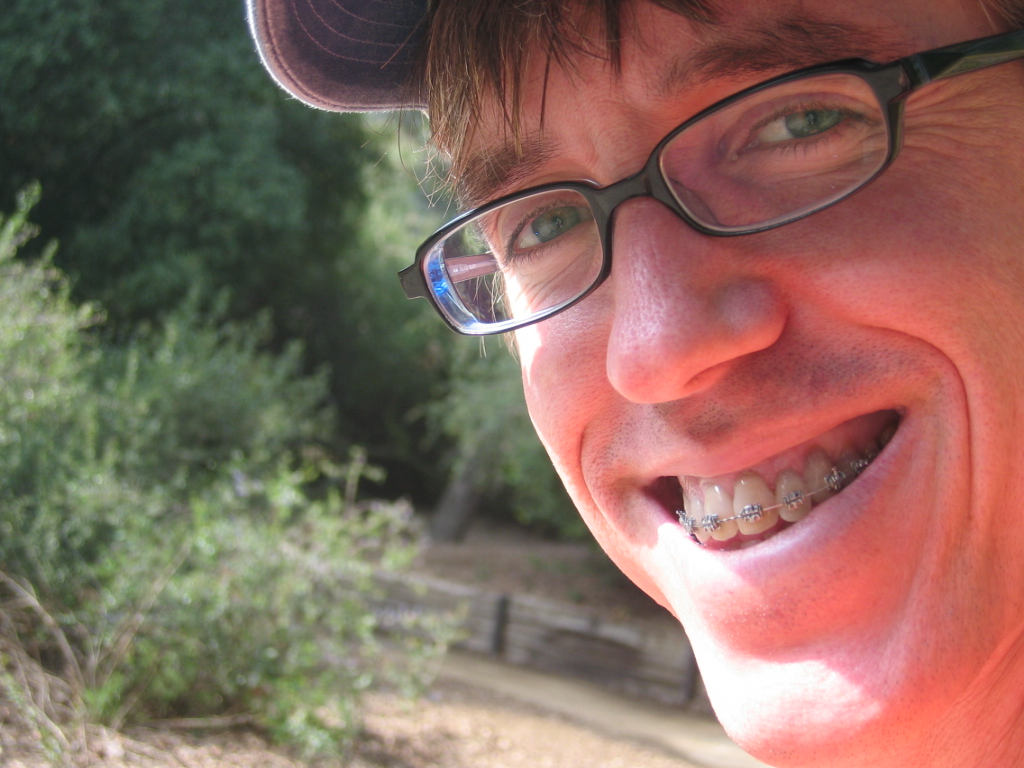smiling man wearing a cap and glasses with trees in the background
