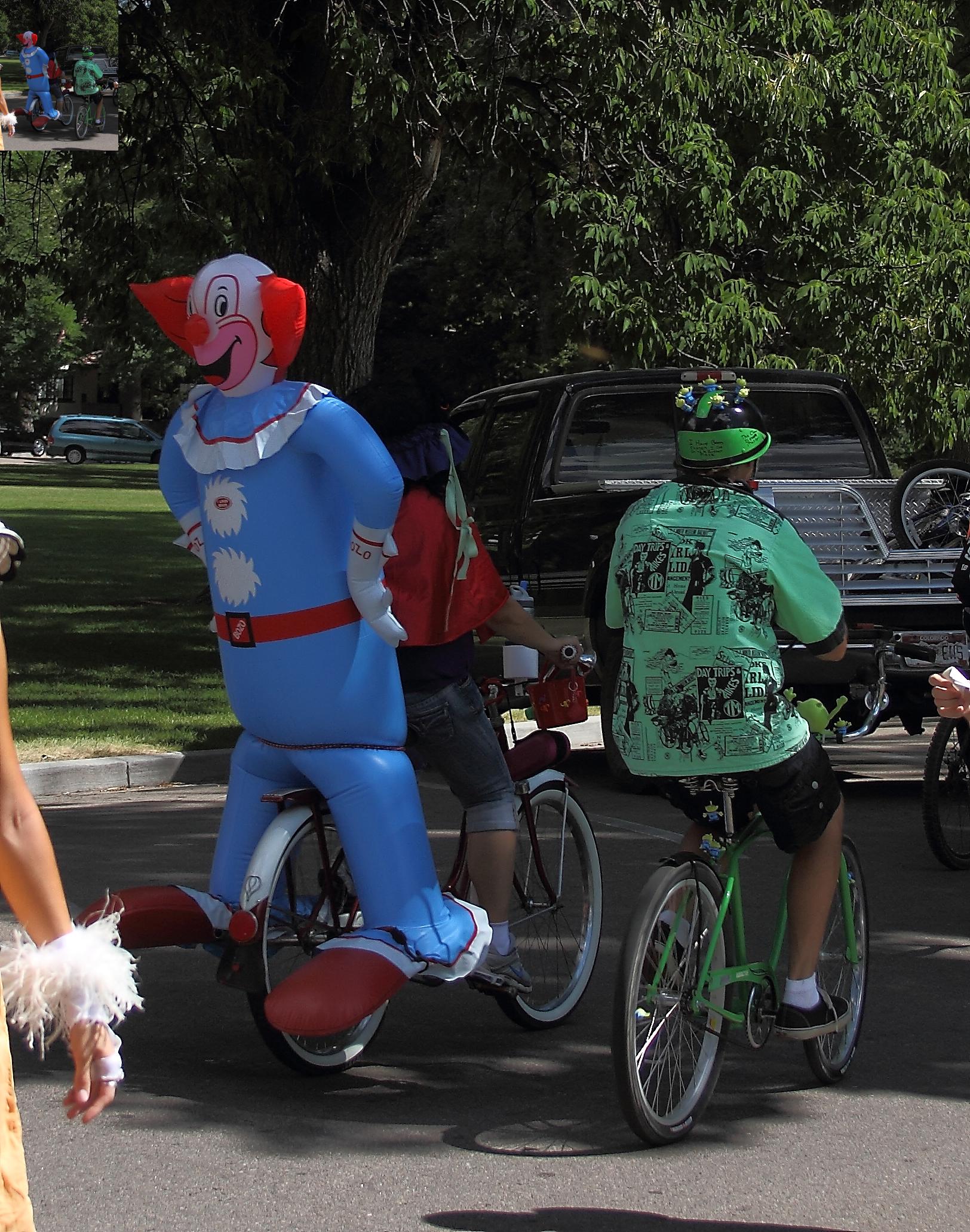 two people in the street, riding on bicycles, with a large balloon man in front of them