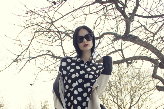 a woman in a black and white polka dotted dress and jacket is standing by a tree with a purse and sunglasses