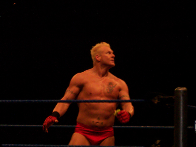 a shirtless wrestling man wearing red trunks holding his hands up in the ring