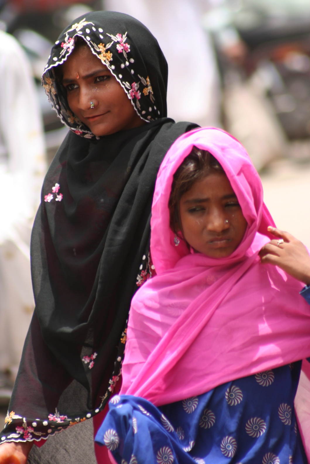 two women in pink scarves and headscarves walk along a busy street