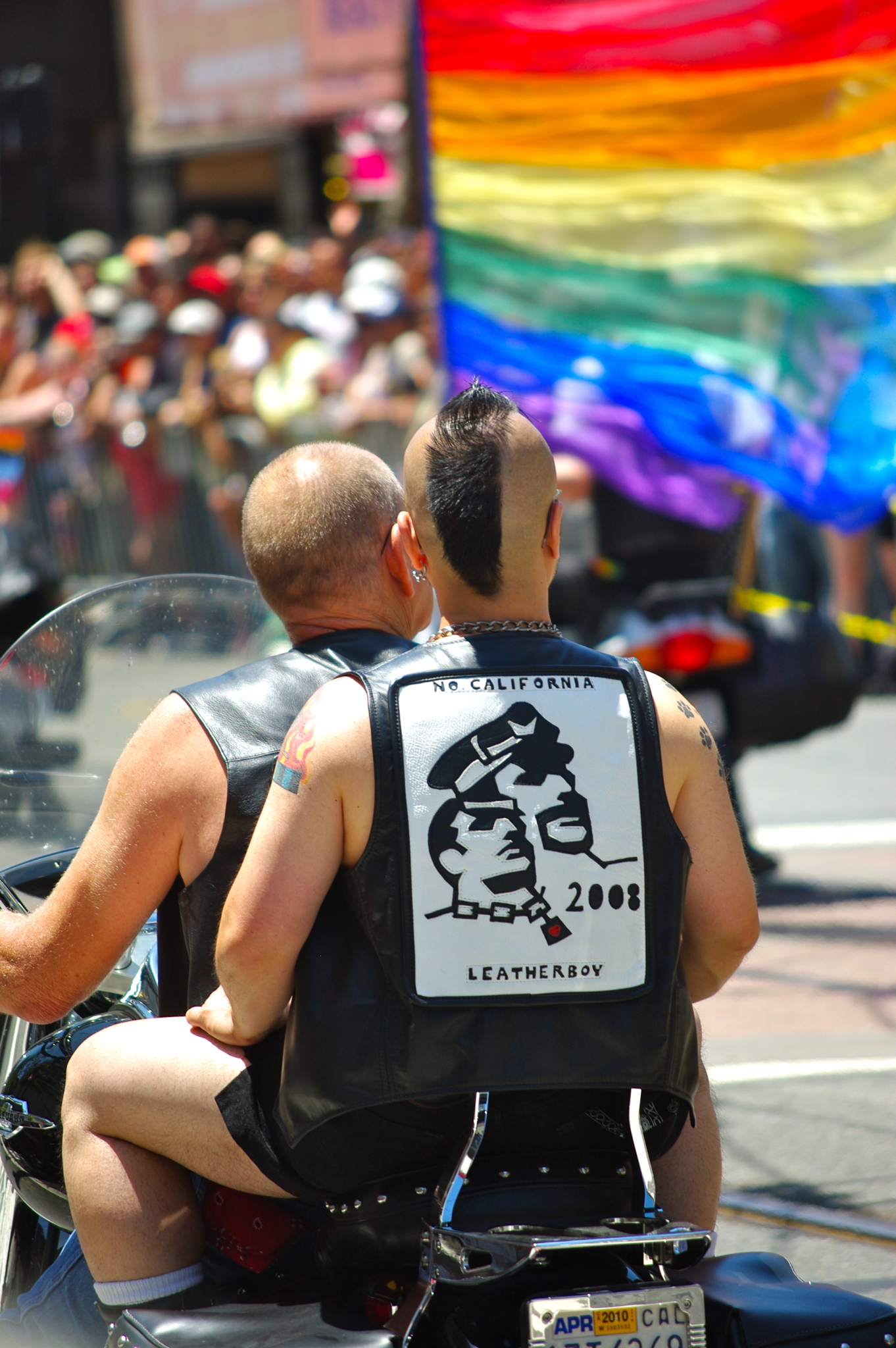 two men on a motorcycle wearing a gay pride vest and one holding the same bike