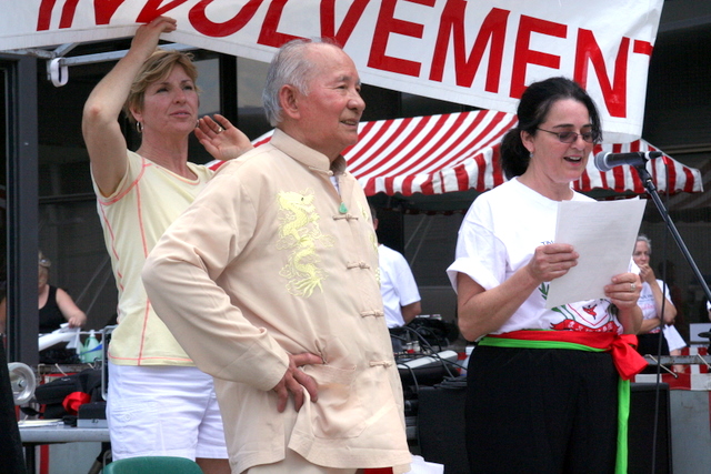 an older man with a white shirt on holds a microphone in one hand and a woman in another stands in front