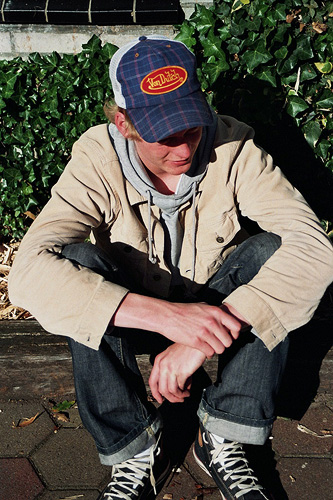 young man in hat sitting on sidewalk with one foot on skateboard
