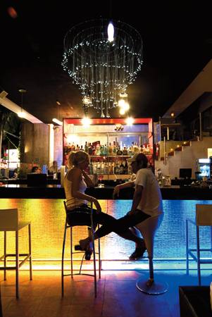 a bar with people sitting at chairs, looking at a pool at night
