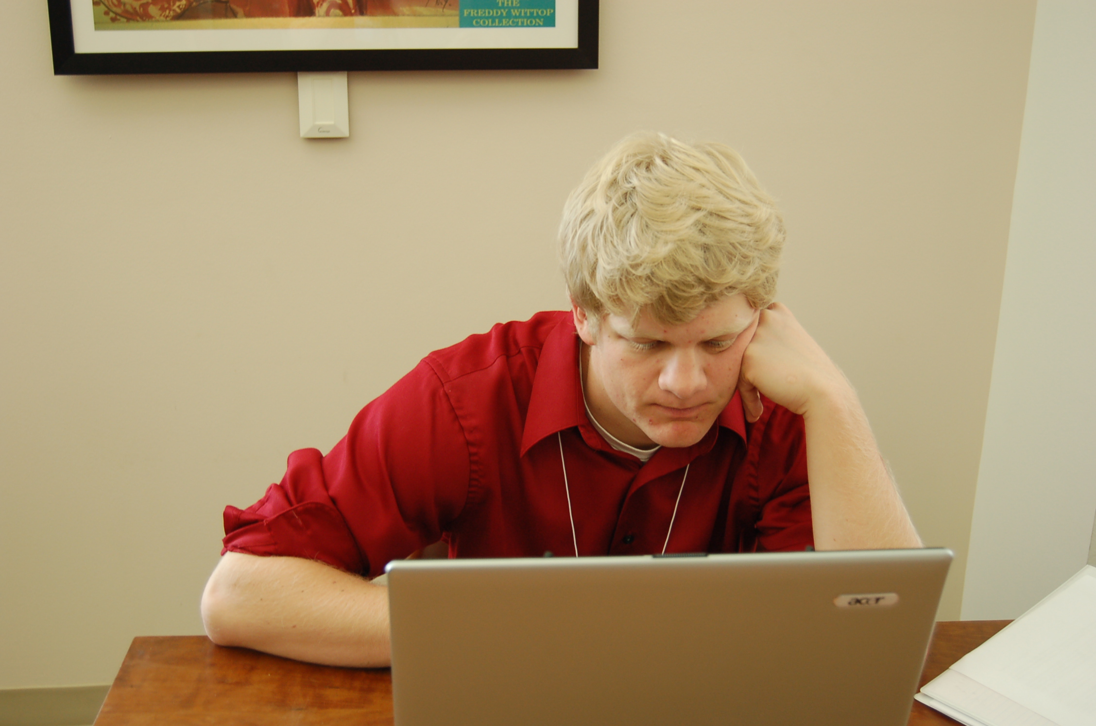 man wearing a red shirt is sitting at a desk looking at his laptop