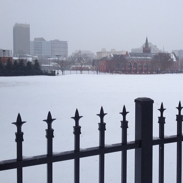 a snowy park has a black fence and city in the distance