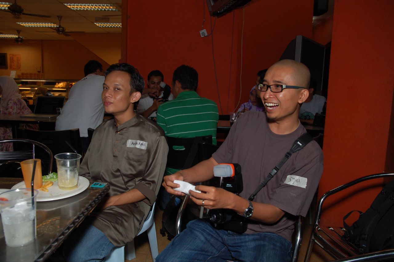 two men sitting at a table while one man holds a wii controller