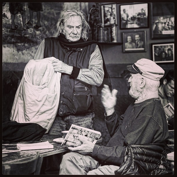 a man wearing a long vest standing over an elderly woman sitting in a chair