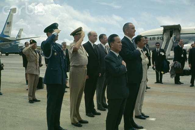 several men in suits and military uniforms stand at an airport while airplanes are parked behind them