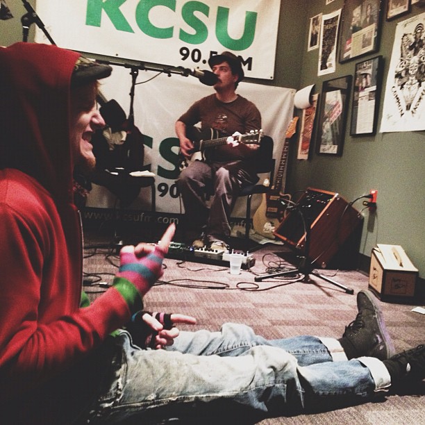 a man on a bed playing guitar and another person wearing a hoodie