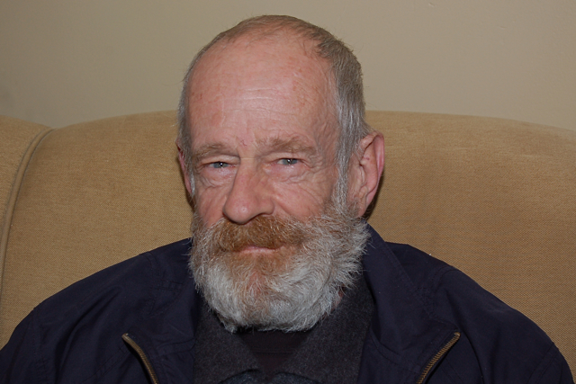an older man with grey hair and a beard sits on a brown sofa