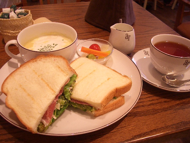 a sandwich sits on a plate with tea, a bowl of soup and another cup