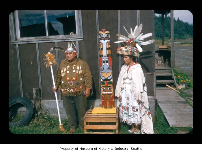 two people in native american outfits stand next to two tall totem poles