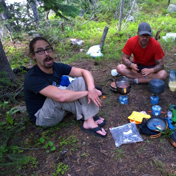 two men sit in the woods and eat camping meals