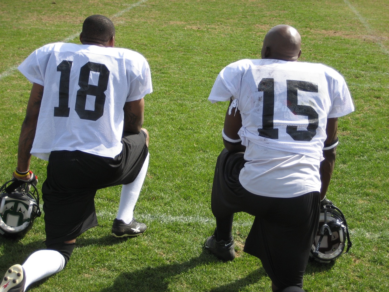 two black men standing on grass while one is holding a helmet