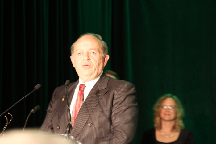 man in business suit talking on stage with two women behind