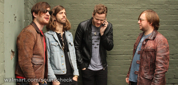 four men standing in front of a brick wall talking on the phone