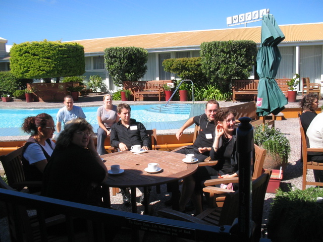 a group of people sit around tables with drinks in front of an outdoor swimming pool