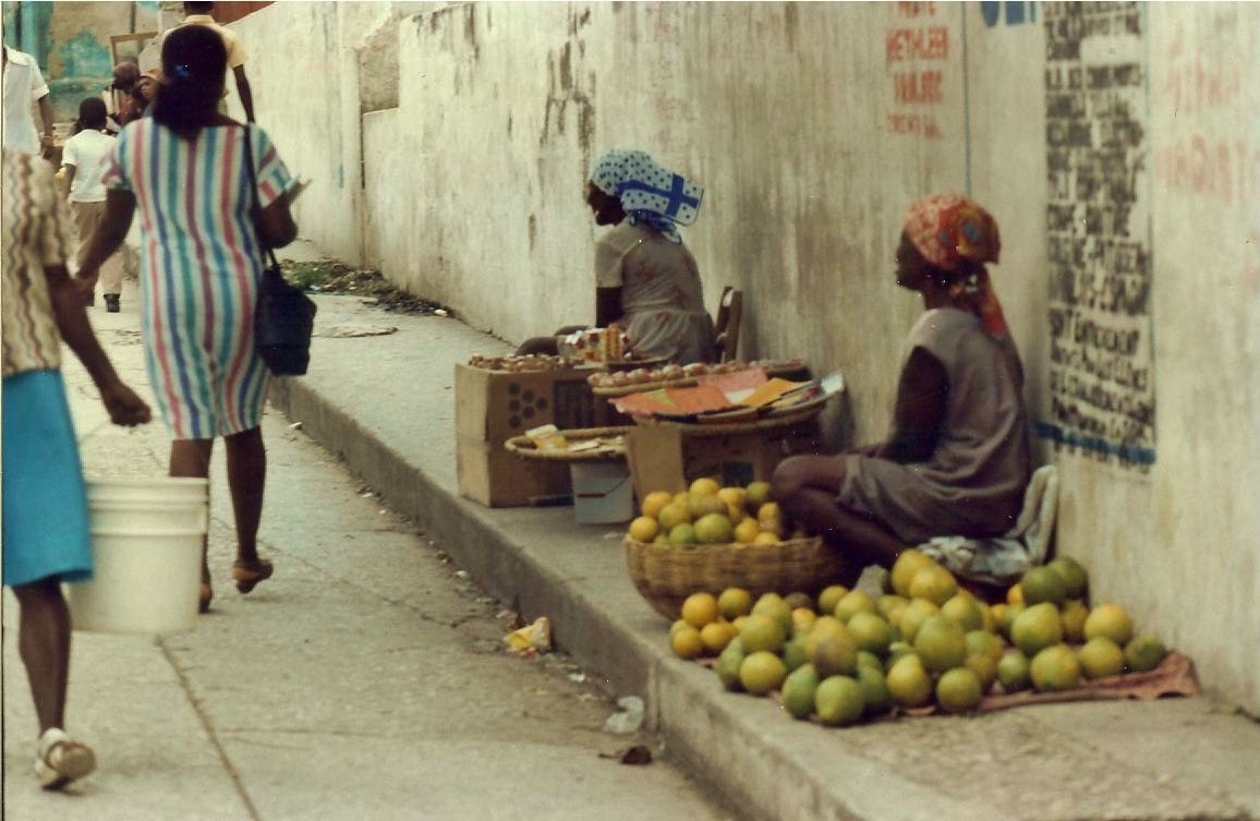 two people sit in front of some fruit while others walk by