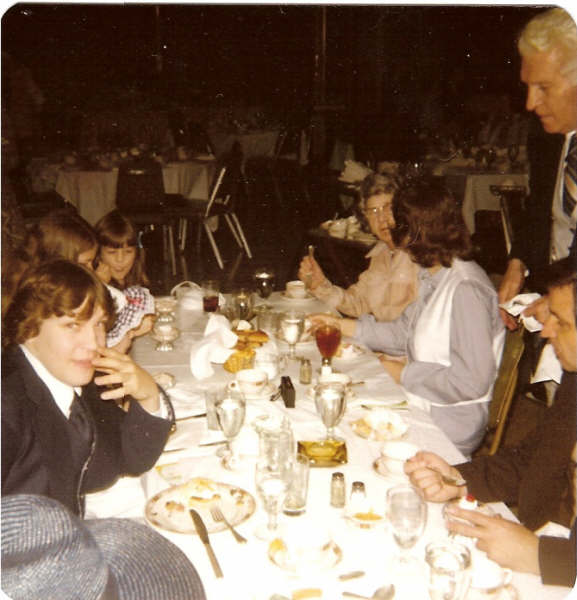 a man stands talking to a group of people seated at a table