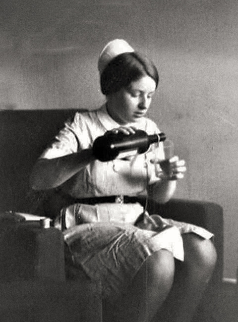 a woman wearing a cap and dress clothes sits on a couch and uses a hairdryer