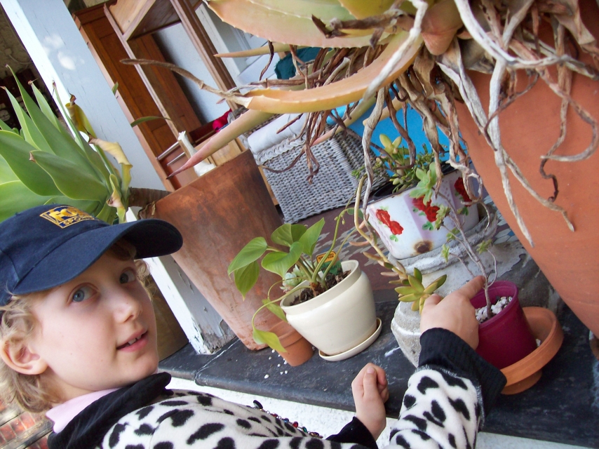 a girl in a blue cap and polka dot top stands next to some plants