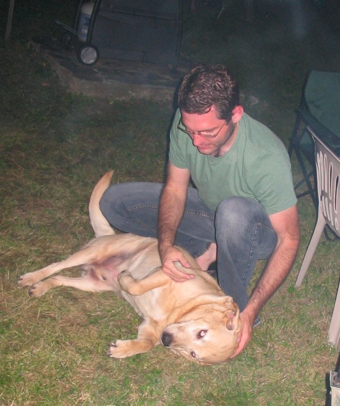a man kneeling down to pet the dog laying on the grass