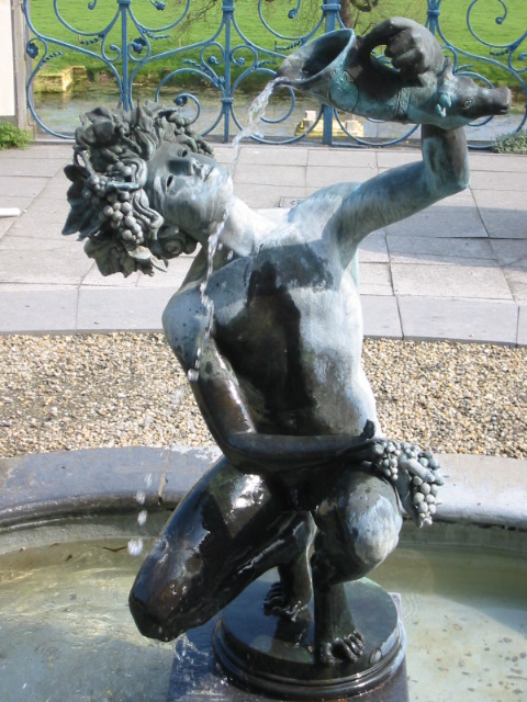 a statue of a man in shorts with a parrot on his shoulder is by a fountain in front of a wrought iron gate