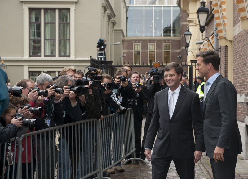two men walk between metal barriers surrounded by press