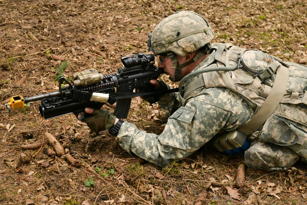 a man in camouflage crouches as he shoots a rifle