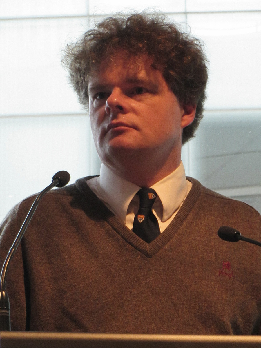 a man wearing a sweater, tie and v - neck shirt is speaking at a podium