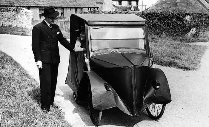 a man wearing a hat, black suit and a tie shaking hands with an antique car