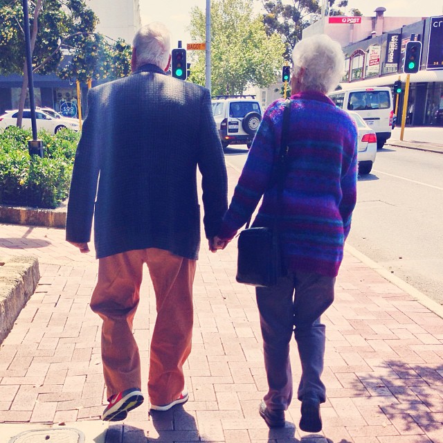 an old man and woman walk on a street holding hands
