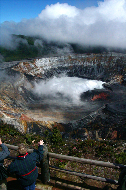 two men taking pictures of the crater while it has steam coming from its boiling water