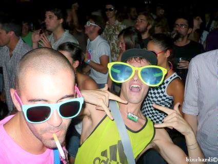 two people with yellow sunglasses with their fingers up and a person holding a straw in the other hand