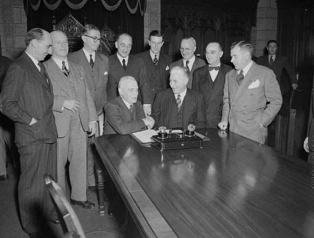 a group of men in suits stand around a wooden table